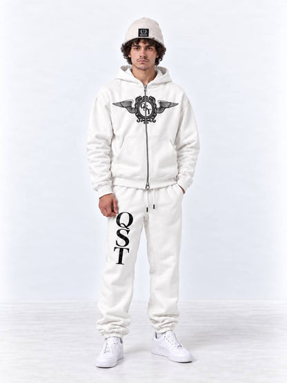 A man modeling a white zip-up hoodie and matching sweatpants set. The hoodie features a black winged crest graphic on the chest, and the sweatpants have 'QST' printed vertically in black on the left leg.